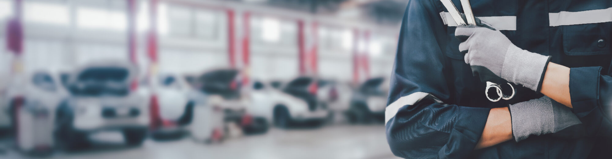 Mechaniker mit verschränkten Armen und Werkzeugen in einer Werkstatt mit mehreren Autos im Hintergrund.