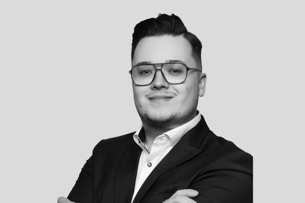 Schwarz-weiß Porträt eines jungen Mannes mit Brille, Anzug und verschränkten Armen.